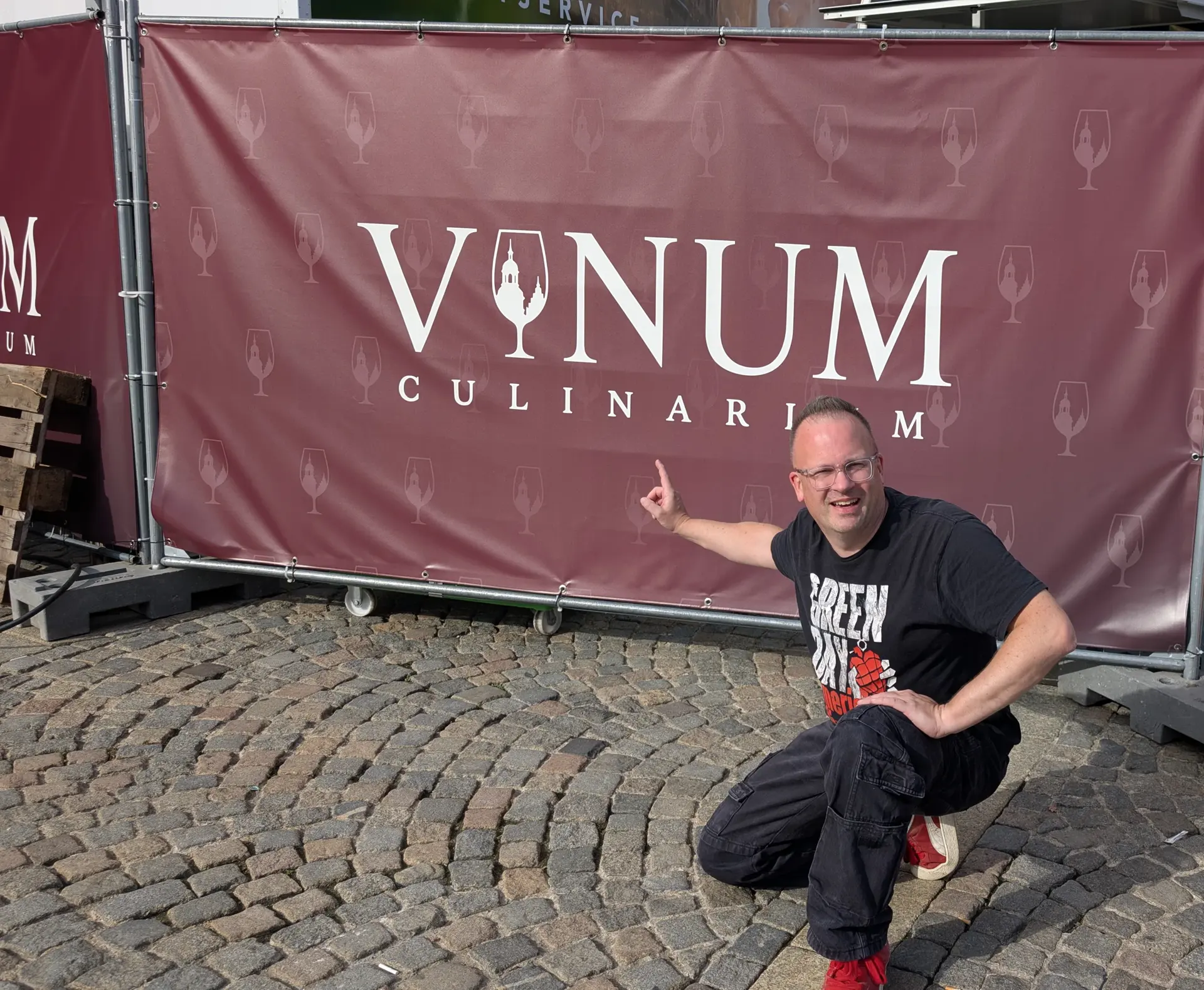 Thomas Peter zeigt auf das Vinum-Culinarium-Bauzaunbanner auf dem Schlossplatz Aschaffenburg